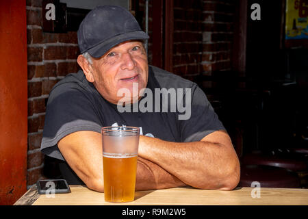 Ein herzhaftes älterer Mann in Schwarz Kappe und T-Shirt genießt ein frisch gezapftes Bier im Fenster eines Huntington Beach, CA, Taverne an einem warmen Sommermorgen. Stockfoto