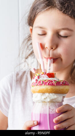 Wenig lächelnde Mädchen Getränke rosa Erdbeere freak shake Cocktail mit Donut und Süßigkeiten, weißen Hintergrund. Ungesunde desserts Konzept. Stockfoto