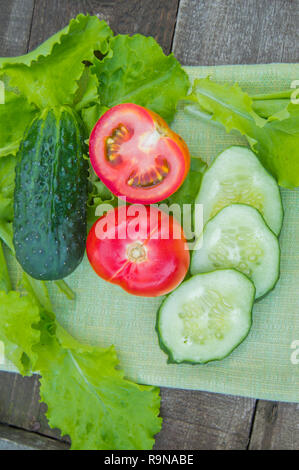 In Scheiben geschnittene Gurken und Tomaten liegen auf einem grünen Serviette, neben Salatblätter, Altes, dunkles Holz- Hintergrund, vertikal gedreht. Stockfoto