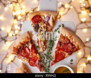 Weihnachten und Neujahr Atmosphäre. Heiße italienische Pizza mit Schmelzen mit Tomate, Salami und Käse auf einem weißen Marmor Schneidebrett. Hintergrund mit Beleuchtung Stockfoto