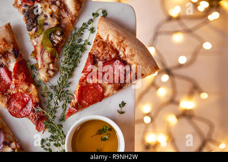 Weihnachten und Neujahr Atmosphäre. Heiße italienische Pizza mit Schmelzen mit Tomate, Salami und Käse auf einem weißen Marmor Schneidebrett. Hintergrund mit Beleuchtung Stockfoto