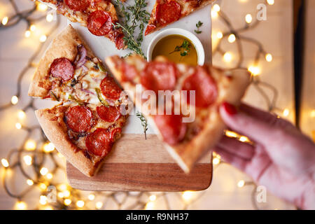 Weihnachten und Neujahr Atmosphäre. Womans hand nimmt Scheibe der Italienischen Pizza mit Schmelzen mit Tomate, Salami und Käse auf einem weißen Marmor Schneidebrett. Stockfoto