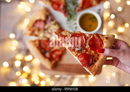Weihnachten und Neujahr Atmosphäre. Womans hand nimmt Scheibe der Italienischen Pizza mit Schmelzen mit Tomate, Salami und Käse auf einem weißen Marmor Schneidebrett. Stockfoto