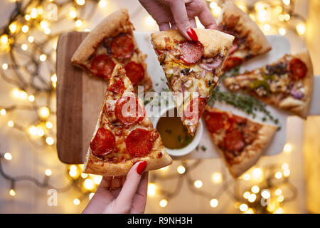 Weihnachten und Neujahr Atmosphäre. Womans hand nimmt Scheibe der Italienischen Pizza mit Schmelzen mit Tomate, Salami und Käse auf einem weißen Marmor Schneidebrett. Stockfoto