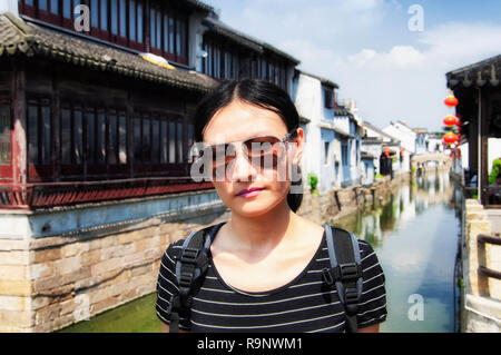 Eine chinesische Frau Sonnenbrille tragen auf einer Brücke mit den Gebäuden der Stadt Luzhi Wasser verschwommen im Hintergrund an einem sonnigen Tag in China. Stockfoto