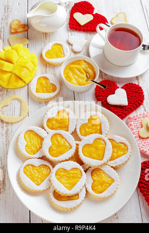 Nahaufnahme der Herzförmigen Linzer Plätzchen mit Mango Marmelade auf einem weißen Teller auf einem Tisch mit gestrickten herzförmige Servietten. eine Tasse Tee, Mango, in Scheiben geschnitten und Stockfoto