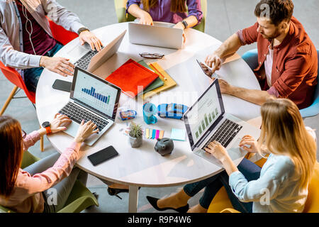 Team von einem jungen Mitarbeiter zusammen mit einigen analitycs auf den Notebooks am runden Tisch sitzen im Büro, Ansicht von oben Stockfoto