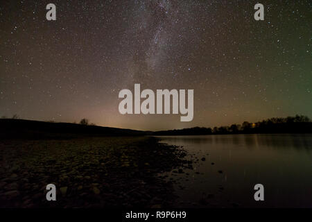 Friedliche Landschaft Panorama in der Nacht. Lange Belichtung geschossen von Kieselsteine Ufer, Bäume am Horizont, helle Sterne und Milchstraße in dunklen Himmel Reflexionseigenschaften Stockfoto