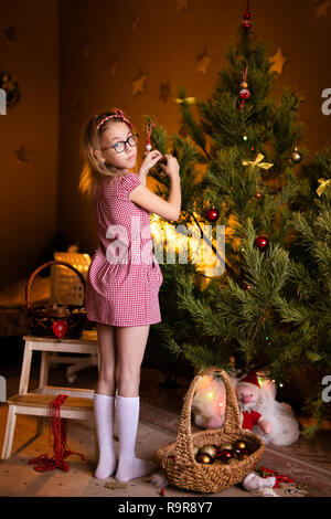 Blonde kind Mädchen im roten Kleid und Kranz mit Gläser Dekoration Weihnachten und Neujahr Baum zu Hause. Stockfoto