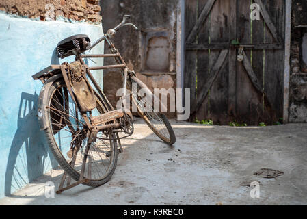 Rostigen alten Fahrrad gegen eine blaue Wand mit Schatten lehnend. Gesperrt Holz- toren im Hintergrund. Bundi, Rajasthan, Indien Stockfoto