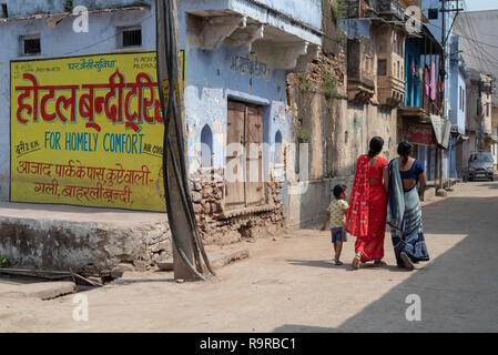 2 indische Frauen in Saris, die von einem kleinen Kind zu Fuß entlang der sonnenbeschienenen Straße mit blau getünchten Gebäuden in Bundi, Rajasthan, Indien gesäumt begleitet Stockfoto