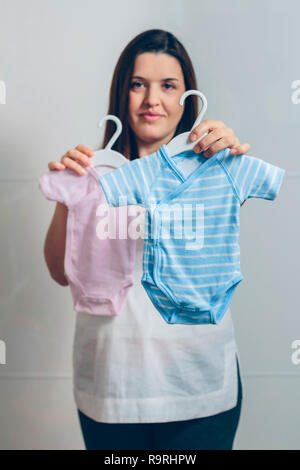 Schwangere Frau mit Baby Bodys indoor. Selektiver Fokus auf Blau Body im Vordergrund Stockfoto