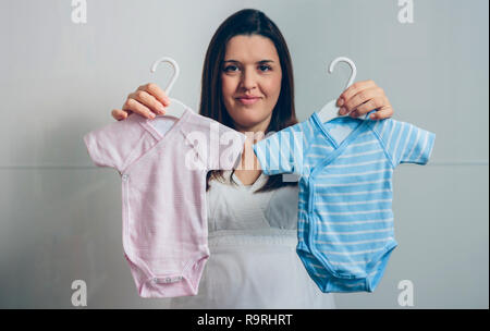 Schwangere Frau, Rosa und Blau Baby Bodys indoor. Selektiver Fokus auf bodysuits im Vordergrund Stockfoto