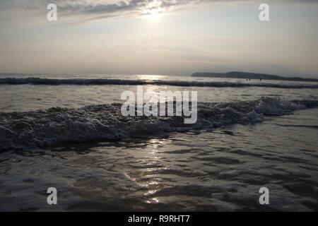 Eine Linie der schäumenden Blasen an einem Sandstrand im Süden Englands Küste, die berühmten weißen Felsen in Silhouette, wie die Sonne und ist reflektiert die Ca Stockfoto