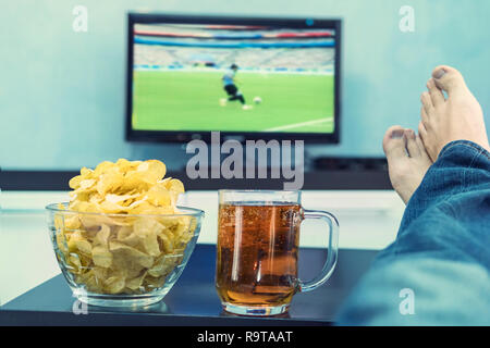 Nahaufnahme der männlichen Hand cup Holding von Bier mit seinen Füßen vor einem großen Fernseher gekreuzt sitzt in seinem Wohnzimmer und beobachtete einen Fuß gesetzt Stockfoto