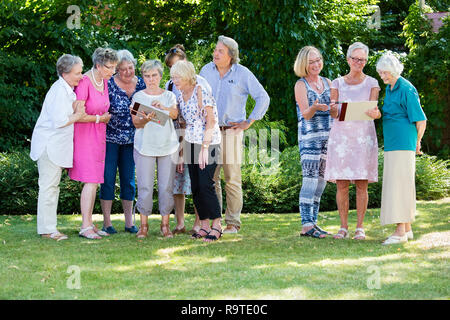 Gruppe von älteren Menschen über ihre Gemälde zusammen mit Kursleiter, beim Stehen im Garten an einem sonnigen Tag. Stockfoto