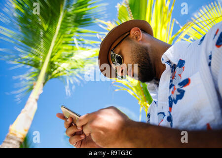 Touristische Mann gekleidet in Geblümten Hemd mit Sonnenbrille und Hut, touristische Mann unter den Kokospalmen während an seinem Handy suchen Stockfoto