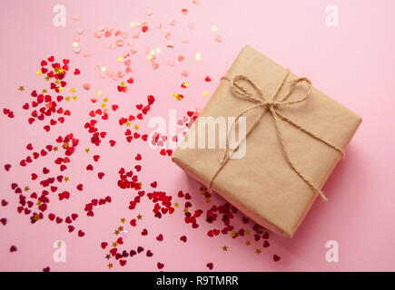 Geschenkbox in braun gefärbte Handwerk Papier gewickelt und gebunden mit Seil auf rosa Hintergrund mit Herzform rot Konfetti. Stockfoto