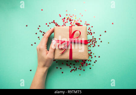 Geschenkbox in braun gefärbte Handwerk Papier gewickelt und mit roter Schleife auf blauem Hintergrund mit rotem Konfetti gebunden. Lady's Hand mit einem geschenkset. Stockfoto