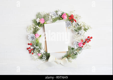 Mockup von Weihnachten Kranz mit Blatt Papier dekoriert mit roten Beeren, Kegel und Rosen. Auf hölzernen Hintergrund Stockfoto