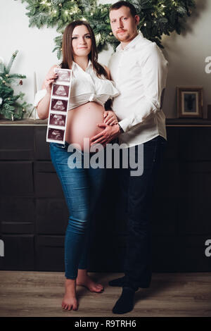 Glücklich der Mann und die Frau zusammen posieren. Ziemlich schwangere Frau mit Ultraschall Bilder der zukünftigen Baby. Mann im weißen Hemd die Hand auf dem Bauch. De Stockfoto