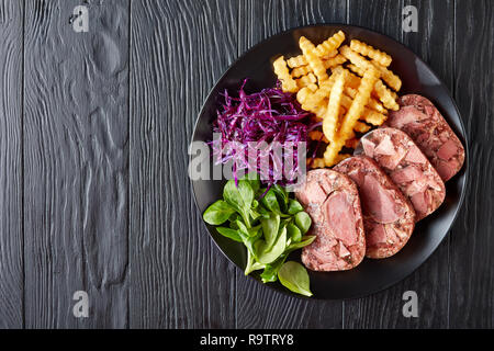 Lecker Geschnetzeltes Rindfleisch Zunge und Fleisch Aspik mit Pommes frites, grüne Blätter und Rotkohl Salat auf einem schwarzen Schild an einem hölzernen Tisch serviert, Ansicht von Stockfoto