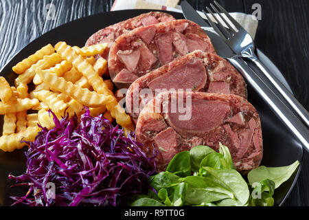 Close-up Geschnetzeltes Rindfleisch Zunge Aspik mit Pommes frites, grüne Blätter und Rotkohl Salat auf einem schwarzen Schild an einem hölzernen Tisch serviert mit Senf in einem Stockfoto