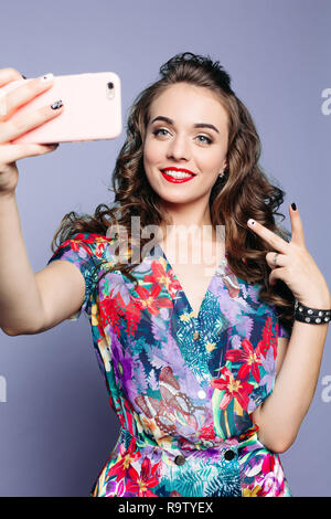 Modische Frau in geblümten Kleid lächelnd und Foto bei Kamera. Brunette Mädchen mit roten Holding Anrufen an Händen und machen selbst portrai Stockfoto