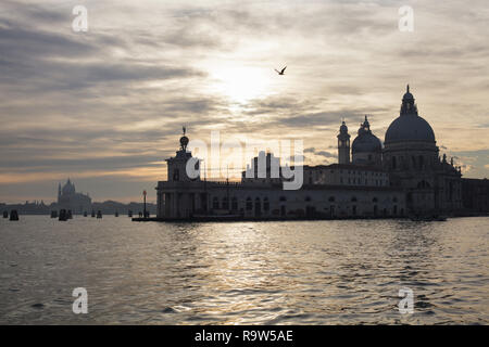 Sonnenuntergang über der Punta della Dogana und die Basilika von Santa Maria della Salute (Basilica di Santa Maria della Salute) in Venedig, Italien. Die redentore Kirche (Chiesa del Santissimo Redentore) auf Giudecca ist im Hintergrund zu sehen. Stockfoto