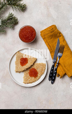 Dünne Pfannkuchen mit roter Kaviar an weiße Platte mit Glas mit rotem Kaviar, Gabel, Messer, Serviette und Tanne in der Nähe von hellen Hintergrund Stockfoto