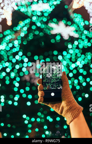 Close-up weibliche Hand in warmen Handschuh holding Handy unter Bild der "grün" leuchtenden Weihnachtsbaum in der Stadt bei Nacht. Stockfoto