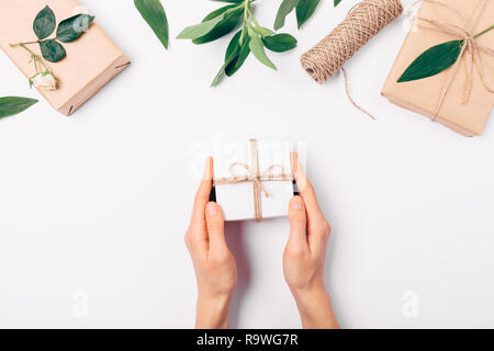 Woman's Hände, die kleine Geschenkbox auf weißen Tisch neben Flach obere Grenze der grüne Blätter, Geschenke und Rose Blume, Ansicht von oben. Stockfoto