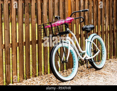 Ein Vintage White Beach Bike lehnt sich an einem braunen Zaun in Ocean Beach, Fire Island, an einem regnerischen Morgen und zwei Schirme in seinem Korb. Stockfoto