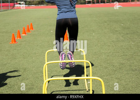 High School Mädchen tun agility Drills über zwei Meter hohen gelben Hürden und orange Kegel auf grünem Rasen Feld während der Leichtathletik Praxis. Stockfoto