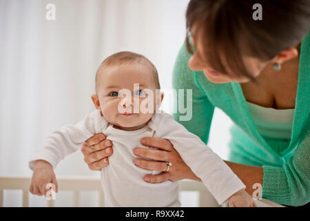 Porträt eines lächelnden baby boy von den Händen seiner Mutter unterstützt werden, als sie ihm hilft, sich in seinem Kinderbett stehen. Stockfoto