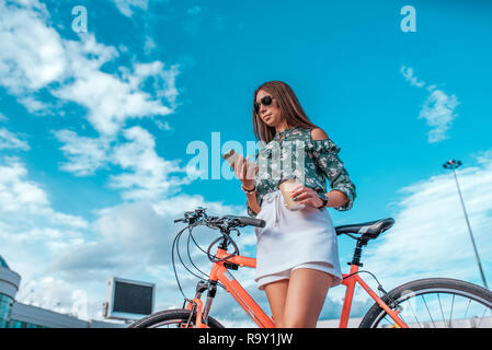 Das Mädchen, das im Sommer in der Stadt, steht neben einer roten Fahrrad, in einem weißen Rock und einem grünen Bluse. In rukaz das Telefon hält, liest die Meldung im Internet. Bei Sonnenbrillen. Stockfoto