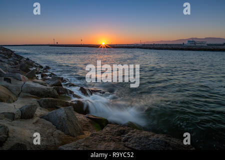 Klar, bunte Himmel mit der Sonne am Horizont bei Sonnenuntergang und Wellen in einem felsigen Jetty, Marina del Rey, Kalifornien Stockfoto