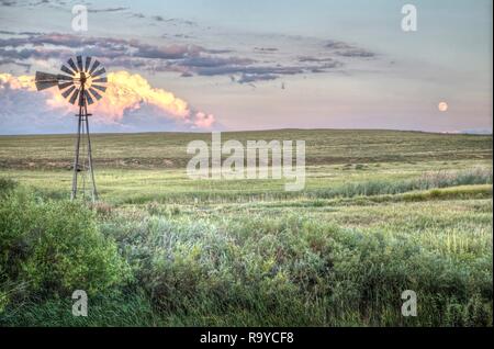 Eine Windmühle auf dem Colorado kurzes Gras Wiese mit einem Hintergrund von Wolken hell durch die untergehende Sonne und den Vollmond. Stockfoto