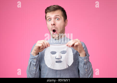 Jungen gutaussehenden Mann Holding kosmetische Maske und Schauen verwundert auf rosa Hintergrund. Stockfoto