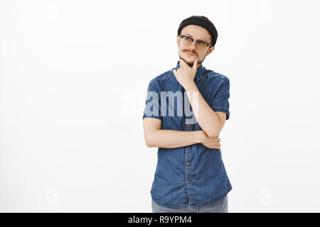 Innen- schuss von nachdenklich attraktive nach kaukasischen Kerl in Schwarz Mütze und Brille reiben Schnurrbart auf Kinn an der oberen, linken Ecke konzentriert Denken, die Entscheidung bzw. Wahl beachten Stockfoto