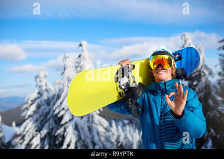 Lächelnd posiert mit Snowboard Snowboarder auf den Schultern am Ski Resort in der Nähe von Forest vor backcountry Freeride und das Tragen von reflektierenden Brille, Farbe Stockfoto