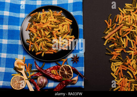 Leckere bunte Italienische Pasta in Keramik Teller mit Löffel aus Holz, Chili, Orange, Knoblauch, Sternanis und Zimt weiß und blau karierten Stoff oder karierten Tuch auf dunklem Hintergrund, Ansicht von oben Stockfoto