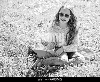 Mädchen sitzt auf Gras an grassplot, grünen Hintergrund. Kind posiert mit Sonnenbrille Pappe ein Lächeln auf den Lippen an der Wiese. Mädchen auf fröhliche Gesicht verbringen Freizeitaktivitäten im Freien. Fashionista Konzept. Stockfoto