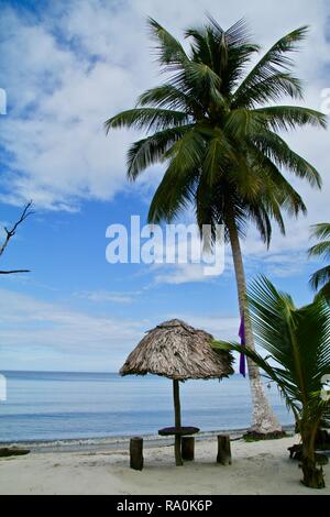 Ein einsamer Tisch und Stühlen unter einem Strohdach Palapa und Palmen am späten Nachmittag an einem karibischen Strand. Stockfoto