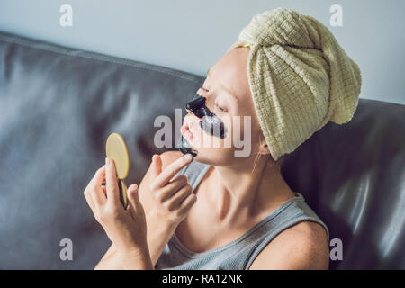 Schöne junge Frau mit Gesichtsmaske zu Hause. Gerne fröhliche Frau, die schwarze Maske auf dem Gesicht Stockfoto