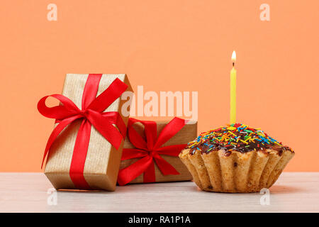 Lecker geburtstag Muffin mit Schokoladenüberzug und Karamell, mit brennenden festliche Kerze und Geschenkboxen auf orange Hintergrund eingerichtet. Happy birthday Mini Stockfoto