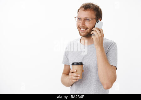 Freundlich Happy Guy Aufruf Freund für morgen Kaffeepause im Cafe holding Smartphone in der Nähe des Ohrs und Pappbecher Lächeln freudig Nette beiläufiges Gespräch stand die Hälfte - Laden links gedreht Stockfoto