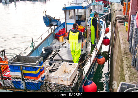 St. Vaast, Normandie, Frankreich - Dezember circa, 2018. Fischer entladen Boxen mit frischem Fang wellhornschnecken Meeresfrüchte aus Fischerboot. St. Vaast und nahe gelegenen Bar Stockfoto