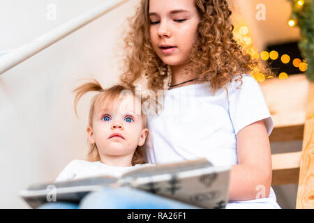 Ältere Schwester lesen Buch vorlesen zu ihrer jüngeren Schwester an der Treppe. Girlande Lichter auf Hintergrund. Familie Liebe und Unterstützung. Stockfoto