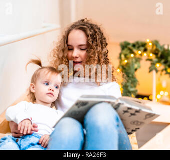 Ältere Schwester lesen Buch vorlesen zu ihrer jüngeren Schwester an der Treppe. Girlande Lichter auf Hintergrund. Familie Liebe und Unterstützung. Stockfoto